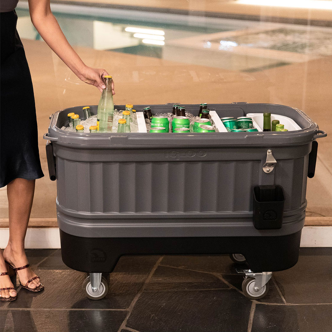 Large wheeled rolling cooler filled with bottled beverages and ice, featuring built-in bottle opener and cup holder for outdoor events and parties
